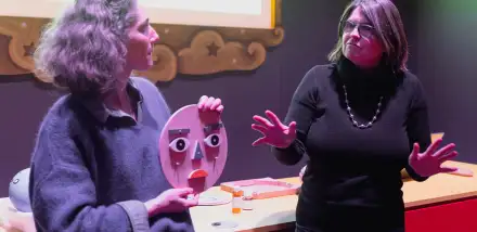 Deux femmes animent un atelier sur les émotions à l'Espace des Sciences. Celle de gauche présente une reproduction en bois d'un visage aux couleurs vives, tandis que l'autre personne donne des explications, les mains grandes ouvertes.