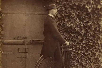 Une photographie en noir et blanc d'un homme en costume. Il se tient de profil sur un  grand bi, une bicyclette qui possède une roue avant d'un très grand diamètre et une roue arrière beaucoup plus petite. Il pose devant une porte et un mur recouvert de lierre.