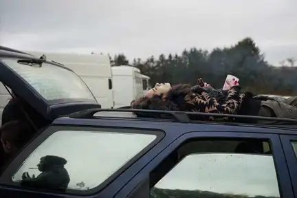 une femme est allongée sur le dos, sur le toit d'une voiture entourée d'autres véhicules. Elle tient dans sa main un gobelet et une bouteille.