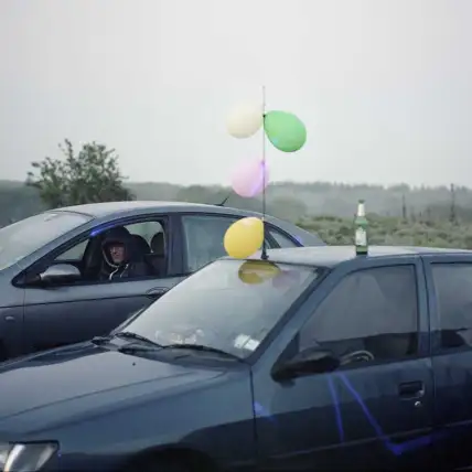 La photographie représente deux véhicules stationnés dans la nature. Le temps est brumeux. La première voiture est décorée avec des ballons de baudruche colorés. Une bouteille est posée sur le toit. Un homme avec une capuche est assis à la place conducteur de la seconde voiture.