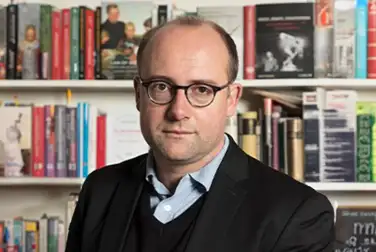 Devant une bibliothèque emplie de livre, Johann Chapoutot regarde face caméra. Il a des lunettes, une chemise bleue et un pull foncé.