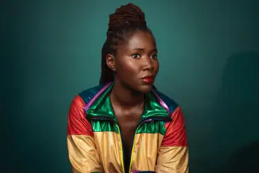 Sur un fond vert dégradé, Alice Diop pose face caméra. Elle regarde hors-champs. Elle porte une veste brillante jaune, verte et rouge.