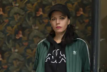 Sur un fond de tapisserie à motif floraux sombre, Laura Vazquez pose face caméra. Elle porte une casquette, des créoles, une veste et un teeshirt de sport.