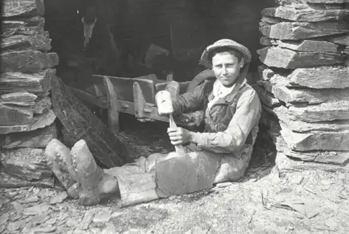 Jeune garçon travaillant devant la porte d'une hutte d'ardoisier, assis sur le sol. Il débite un morceau d'ardoise. A l'intérieur de la hutte, un homme plus agé travaille également, une brouette bloque l'entrée.