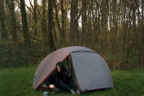 Une tente de tente sur une pelouse devant un bois. Une personne est assise dans l'embrasure, tournée vers le réchaud qu'elle bricole.