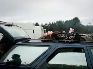 Sur le toit d'une voiture, en manteau de fourrure, rouge à lèvre rouge et gobelet plastique à la main, une jeune femme regarde le ciel.