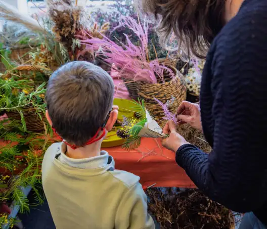 Sur une table recouverte de tissus, un enfant et une femme de dos font de petits bouquets de fleurs séchées dans les cornets de papier journal.