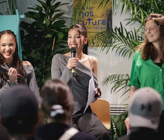 Sur une scènes, trois jeunes femmes parlent et rient face au public.