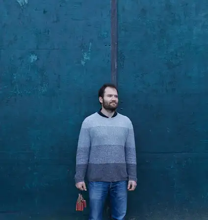 Devant une porte en métal bleu, en bottes dans la boue, un homme brun avec une barbe et un pull bleu souris légèrement en regardant à droite.