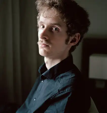 De biais, un jeune homme à moustache et à la mine boudeuse regarde la caméra.