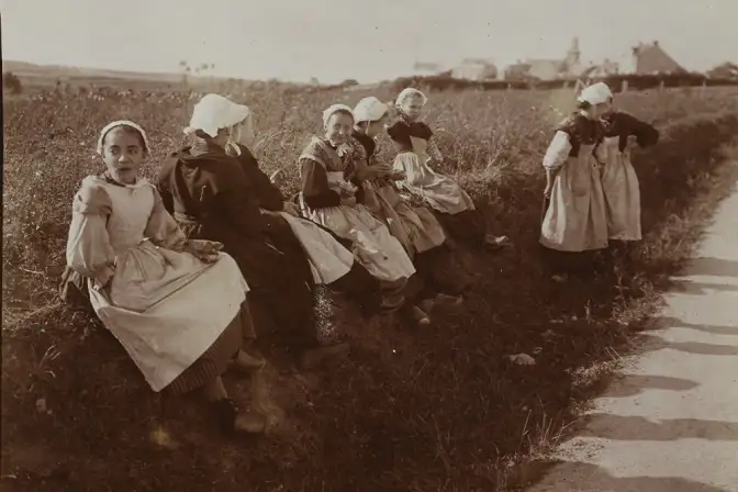 Prise de vue en sépai, le long d'un chemin : plusieurs jeunes filles en tablier et coiffes sont assises sur un talu.