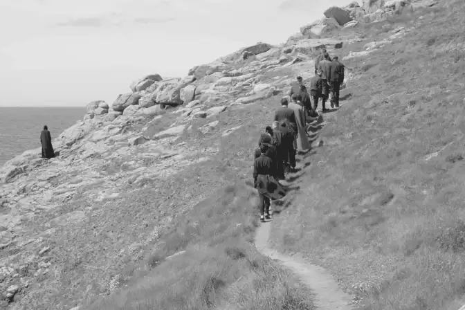 Cette photographie, prise près d'une côte maritime, nous montre 16 personnes vêtues de costumes d'époque en Bretagne. Elles marchent sur un petit chemin tracé au bord de la côte. Cependant, une personne est en dehors du chemin, très proche du précipice qui marque la fin de la côte.