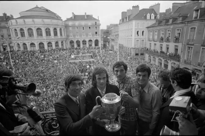 Au premier plan six joueurs du stade rennais qui tiennent la coupe, depuis le balcon de l'hôtel de ville. derrière eux une foule de supporters sur la place de la mairie