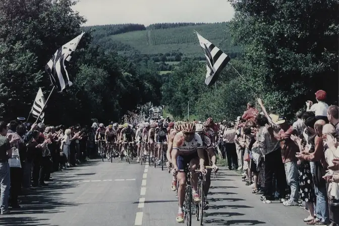 Le peloton du tour de France progresse sur une route de Bretagne, il est entouré de public au bord de la route avec plusieurs drapeaus bretons