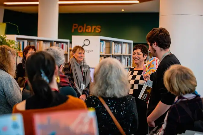 Dans la Bibliothèque, une autrice déambule, accompagnée de personnes l'écoutant. Elle sourit.