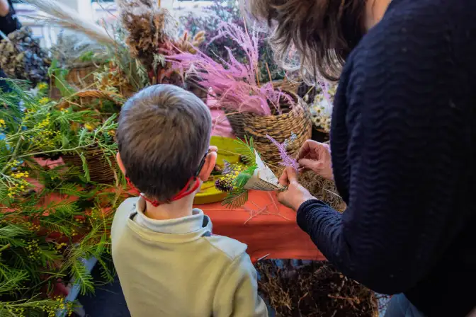 Sur une table recouverte de tissus, un enfant et une femme de dos font de petits bouquets de fleurs séchées dans les cornets de papier journal.