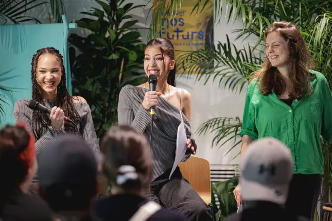 Cette photo présente trois femmes en train de présenter une idée à un public lors des éditions Nos Futurs. Elles sont en train de rigoler, micro en main, face à un public que nous voyons de dos.