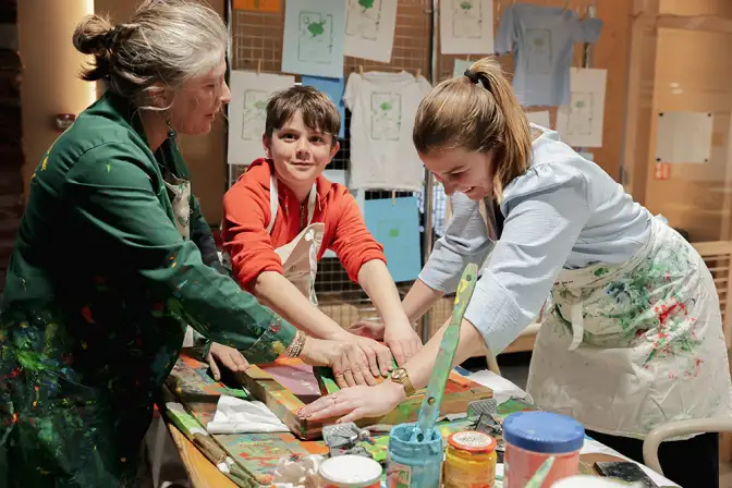 Cette photo nous montre un atelier de peinture au pochoir sur t-shirt, à l'occasion de l'événement Nos Futurs 2024. On y perçoit deux femmes et un jeune garçon. L'une d'elles tient le pochoir au-dessus du t-shirt, tandis que l'autre femme et le garçon appliquent la peinture.