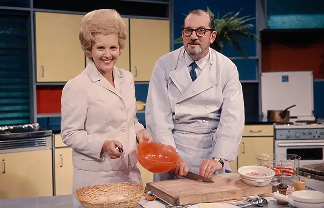 Dans une cuisine des années 60, deux personnes, une jeune femme et un home d'âge mur à lunettes, en blouse blanche, sourient à l'objectif. Devant eux, une planche à découper, un panier en osier, un saladier en plastique orange, et divers objets de cuisine. La photo est légèrement humoristique du fait de la coiffure impressionnante de la femme : un "beehive" blond assez haut.