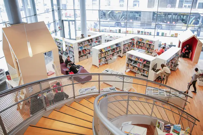 La photo est prise en haut d'un escalier en colimaçon léger. On y voit des rayons de livres blancs, un sol en parquet clair, deux structure de jeux en forme de cabane minimaliste en bois très clair.