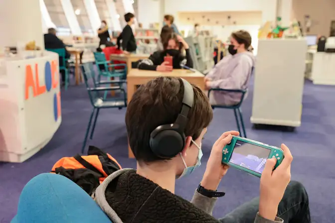 Au premier plan, on voit un jeune garçon de trois -quart, aux cheveux court brun, avec un casque glissé derrière l'une de ses oreilles, avec un masque (on est encre en plein covid sur cette photo). Il joue à la Switch, assis sur un fauteuil bleu turquoise. Au second plan, on distingue des tables en bois clair, des chaises en métal  turquoise et en bois clair, des étagères blanches dans lesquelles sont entreposés les livres, et un sol en moquette bleue marine.