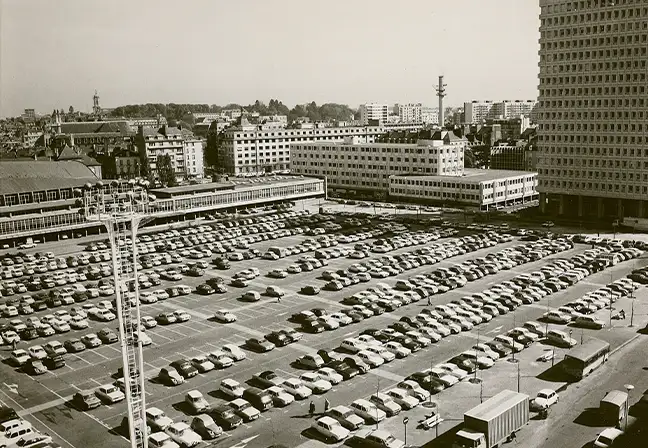 Photo en noir et blanc. L'actuelle place piétonne Charles de Gaulle est encore le parking du Champs de Mars sur cette photo. C'est un immense parking à ciel ouvert. On distingue déjà la tour de la CAF, le bâtiment du Liberté, mais pas encore le cinéma du Gaumont.