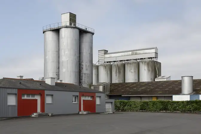 Une photographie d'un paysage industriel : deux bâtiments gris coupent l'image de manière horizontale. Au centre,  en arrière plan, des blocsde silos de taille différentes se dressent.  Le ciel est bleu pâle, et quelques touchent de couleur éclairent la scène : une haie verte le long d'un des bâtiment, et les portes rouges de l'autre.