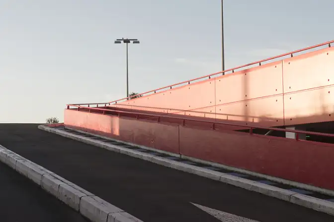 Prise de vue du dernier étage d'un parking, depuis la route en bitume noir qui grimpe en direction du toit. On distingue une flèche blanche au sol, et une rampe d'accès rouge dans une lumière pâle, matinale. Le ciel est d'un bleu pâle qui contraste avec le sol sombre et la rampe rouge. Derrière la rambarde rouge délimitant les contours du parking s'élèvent deux grands lampadaires.