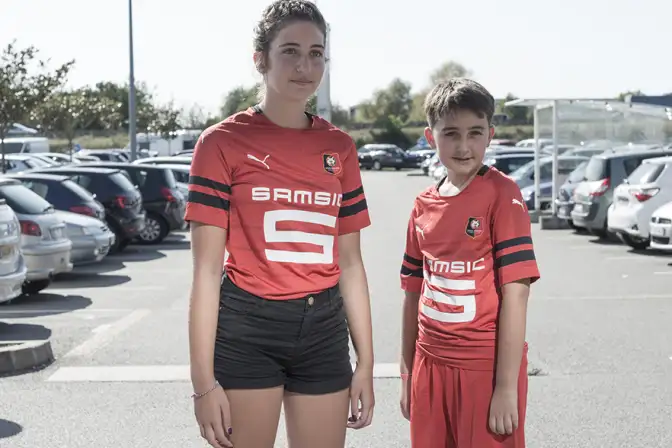 Sur un parking, au soleil, encadrés de rangées de voitures, deux jeunes supporters du stade rennais regardent à droite, plus loin que l'objectif. On les image frère et soeur : l'une, plus grande, les cheveux bruns attachés, porte un short de jean foncé et un mailleur rouge aux quelques lignes de noir et aux inscriptions blanches "SAMSIC". Le second, plus petit, souris doucement. Il est habillé avec le même maillot, plus un short rouge assorti. Il poste les cheveux court du même brun que la première, et ses traits portent encore la rondeur de l'enfance.