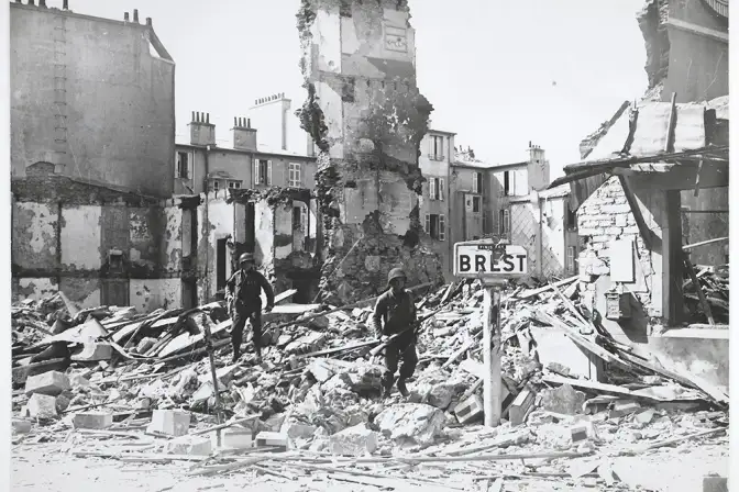 Photographie prise au moment de la libération de Brest en septembre 1944 ; deux soldats américains se déplacent sur les ruines de maisons ; au premier plan, un panneau indicateur routier avec "Finistère / BREST".