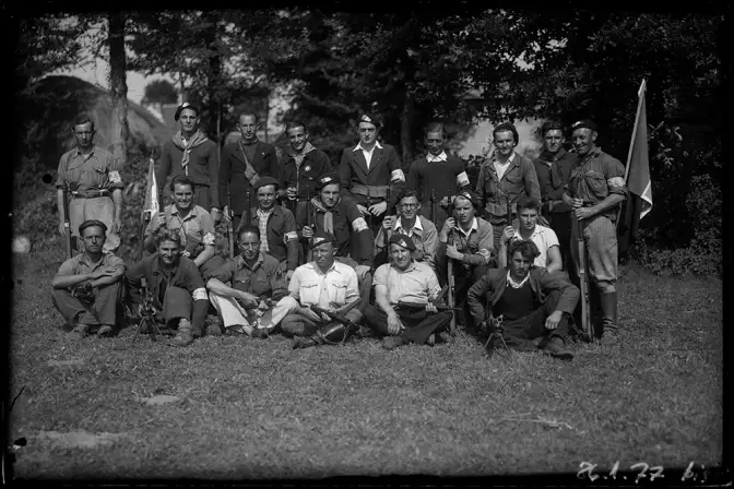 Groupe de résistants de la région de Saint-Nicolas-du-Pelem pris dans un champ. Sur trois rangs : au premier rang, les hommes assis avec leurs mitraillettes ; au second rang, à genoux avec leurs fusils ; dernier rang, debout avec drapeau et fanion à droite et à gauche. Certains portent le brassard.