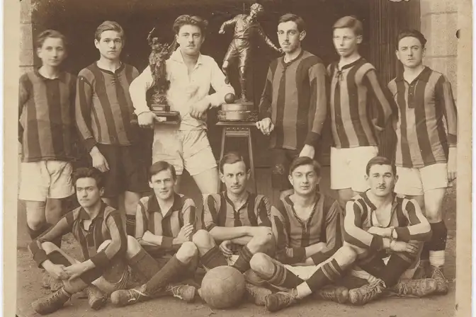 Portrait de groupe de l'équipe de foot-ball Saint-Martin. Les lycéens posent en tenue de sport avec, au centre, les deux trophées remportés lors de la compétition. Le premier représente un joueur de football, le second représente deux femmes dont une ailée. Aux pieds des sportifs de la première rangée, on aperçoit un ballon.