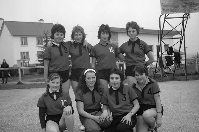 Équipe de basket féminine sur le terrain municipal de Saint-Nicolas-du-Pelem. 8 jeunes filles en maillot sur un sol en bitume clair, en extérieur. Elles sourient.