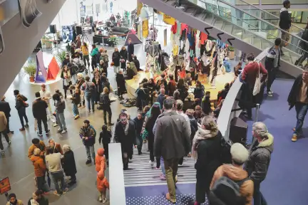 La photo est prise depuis l'escalier de la coursive, ofrrant une vue en contre-plongée sur le hall des Champs Libres. De nombreuses personnes descendent et déambulent dans l'espace.