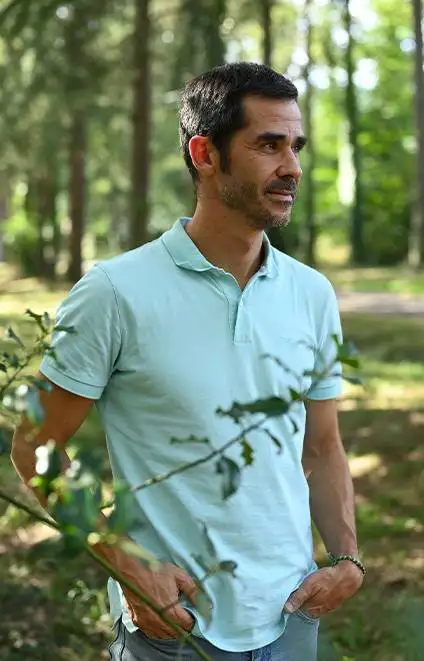 Une photographie d'Anthony Berthou en chemise bleu clair, de trois-quart. Il regarde au loin, entouré de végétation.