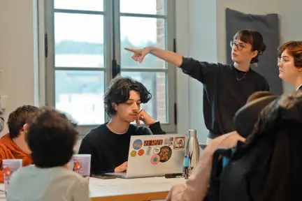 Photo prise lors du hackathon pour l’événement Nos Futurs. On y voit une jeune femme debout, pointant quelque chose du doigt. À côté d’elle, de jeunes gens sont assis à un bureau, semblant concentrés et attentifs.