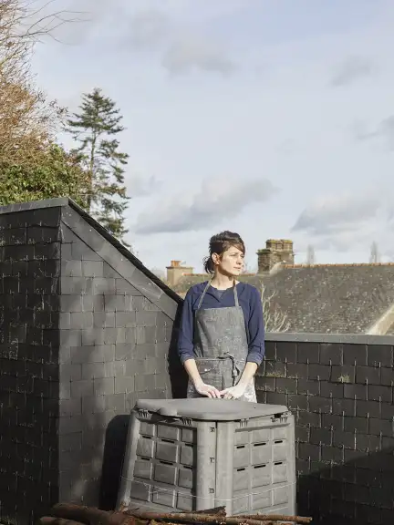 Sur un toit d'ardoises, devant un grand contenaire en plastique noir, se tient une femme en tablier. Elle tient ses mains couvertes d'argile blanche devant elle et regarde à droite de la photo avec un air contemplatif, les lèvres un  peu serrées. Elle a les cheveux attachés en chignon bas. Derrière elle, on distingue d'autre toits, quelques cîmes d'arbres, et un ciel bleu clair.