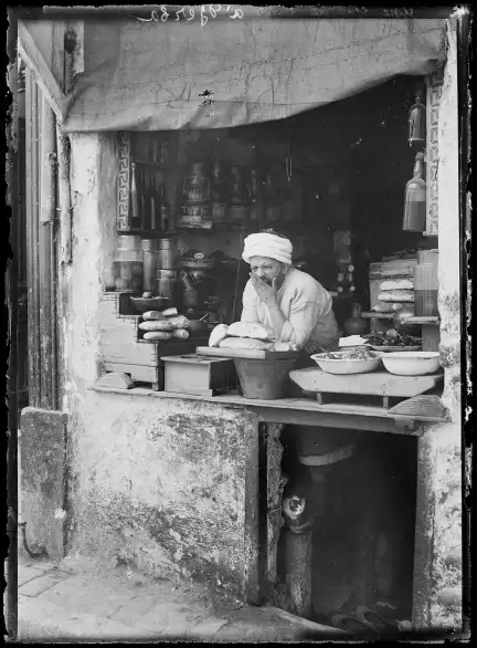 Vue d'un marchand dans sa boutique. Coiffé d'un turban, il baille et porte la main devant sa bouche. Au-dessus du comptoir, une bâche (pour fermer la boutique). À l'intérieur, des jarres, des récipients et des bouteilles sont sur des étagères. Sur le comptoir, des pains ; à droite, des préparations culinaires. Sous le comptoir, une ouverture carrée : on aperçoit un chaton et une paire de chaussures.