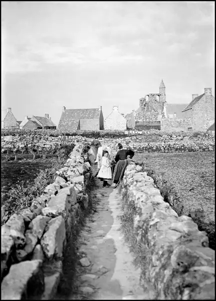 alt="Vue de deux femmes, d'une fillette et d'une vache entre deux murets de pierre. À l'arrière-plan, le village de Kérity Penmarc'h et l'église Sainte-Thumette."