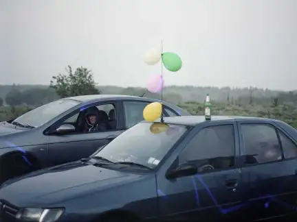 La photographie représente deux véhicules stationnés dans la nature. Le temps est brumeux. La première voiture est décorée avec des ballons de baudruche colorés. Une bouteille est posée sur le toit. Un homme avec une capuche est assis à la place conducteur de la seconde voiture.