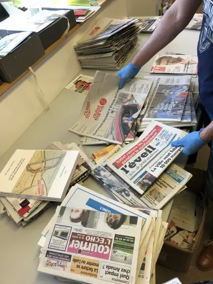 Cette image nous montre plusieurs journaux en train d'être triés par un homme portant des gants en latex bleu. On perçoit différents livres, magazines et journaux tels que Ouest France.