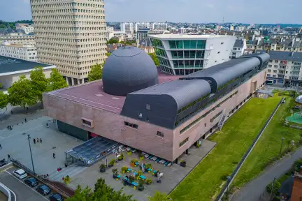 Vue en contre-plongé du bâtiment des Champs Libres : on y voit le Musée avec ses grandes plaques de schiste rouge, la tour blanche vitrée de la Bibliothèque, et le dôme en tuiles d'ardoise noire du Planétarium. On y voit également la terrasse du Café avec ses petits arbres.