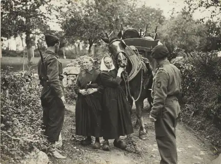 Photo en noir et blanc légèrement jaunie. Sur un petit chemin de parc aménagé, deux femmes vieillissantes habillées en robes noir et portant la coiffe du pays de Lorient tiennent pour l'une la bride d'un lourd cheval de trait qui mène une carioles de chaises, foin et autres objets emportés dans la fuite, et l'autre s'adresse en souriant légèrement à deux combattants des F.F.I.