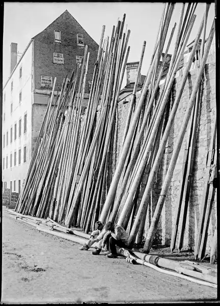 Vue de deux pécheurs, en béret et en sabots, se reposant, assis sur un mât. Derrière eux, plusieurs dizaines de mâts sont légèrement inclinés sur un grand mur de pierre.