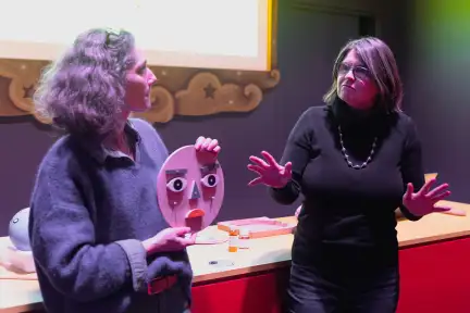 Deux femmes animent un atelier sur les émotions à l'Espace des Sciences. Celle de gauche présente une reproduction en bois d'un visage aux couleurs vives, tandis que l'autre personne donne des explications, les mains grandes ouvertes.