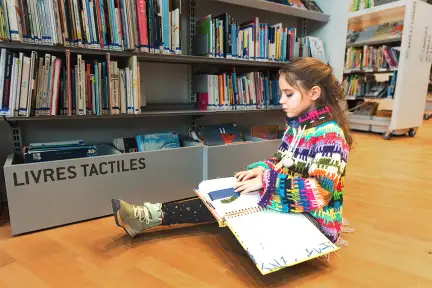 Une fillette brune au gilet multicolore,  lit un livre en braille et relief avec ses doigts. Elle est assise parre-terre près du bac des livres tactiles, Chez les Enfants à Bibliothèque.