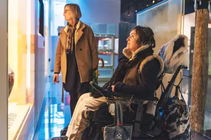 Deux femmes dont l'une en fauteuil, regardent une vitrine bien éclairée du Musée de Bretagne. On y voit des fragements de poterie.