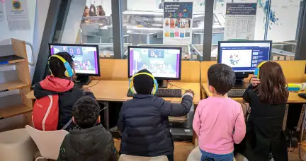 Sur cette image, on peut voir cinq enfants dans le rez-de-chaussée de la Bibliothèque. Trois d'entre eux sont assis devant des ordinateurs, concentrés sur leurs jeux, avec des casques sur les oreilles. Les deux autres enfants se tiennent à côté, observant les jeux de leurs camarades.