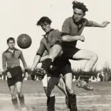 Une photographie en noir et blanc d'un match de football. L'un des joueurs se tient en l'air alors que sa jambe vient de taper dans le ballon.