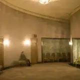 Dans un long couloir en arc de cercle, éclairé de lumières électrique, avec de nombreuses chaises alignés sur les côtés, un homme est assis et regarde vers le fonde de la pièce.